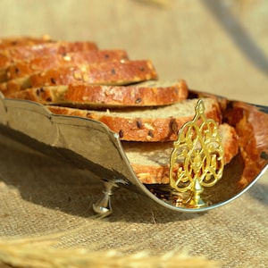 Puede incluir: Una cesta de pan de plata con un asa decorativa dorada, llena de rebanadas de pan multigrano. La cesta está sobre una superficie de arpillera.