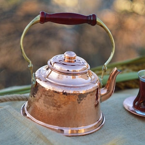 Puede incluir: Una tetera de cobre martillado con un asa de madera y un pico de latón. La tetera está sentada sobre una superficie de arpillera.