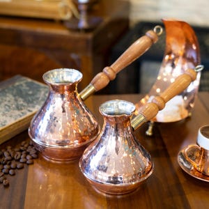 Puede incluir: Dos cafeteras de cobre con mangos de madera sobre una superficie de madera, junto a granos de café y un libro antiguo. Un calentador de café de cobre y una taza de café también están presentes, sugiriendo un ambiente de café.