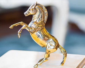 Puede incluir: Una figurita de caballo de metal dorado de pie sobre sus patas traseras con la cabeza en alto. La figurita está sobre una superficie de color claro.