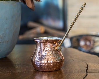 Pode incluir: Uma cafeteira de cobre com acabamento martelado e uma alça longa e adornada. A cafeteira está sobre uma superfície de madeira.