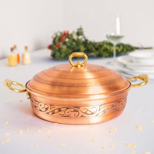 Könnte beinhalten: Ein Kupfertopf mit Deckel und zwei Griffen. Der Topf hat ein verziertes Design am Rand und steht auf einer weißen Oberfläche.