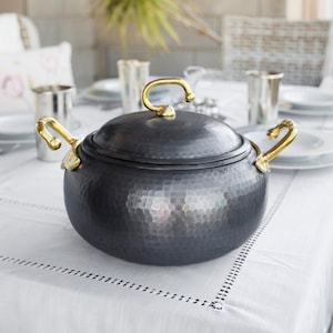 May include: A hammered black metal pot with gold handles and a lid. The pot is sitting on a white tablecloth.