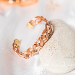 Op de afbeelding: Een koperen draad armband met een gevlochten ontwerp. De armband ligt op een witte steen.
