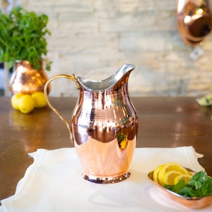 May include: A copper pitcher with a silver rim and a gold-colored handle sits on a white cloth. Sliced lemons and mint leaves are in a copper dish. Two copper pots hang in the background, suggesting a kitchen setting.