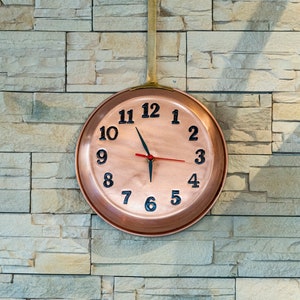May include: A copper-coloured wall clock shaped like a frying pan, with black numerals and red hands. The clock is mounted on a light-coloured stone wall. The handle is brass-coloured.