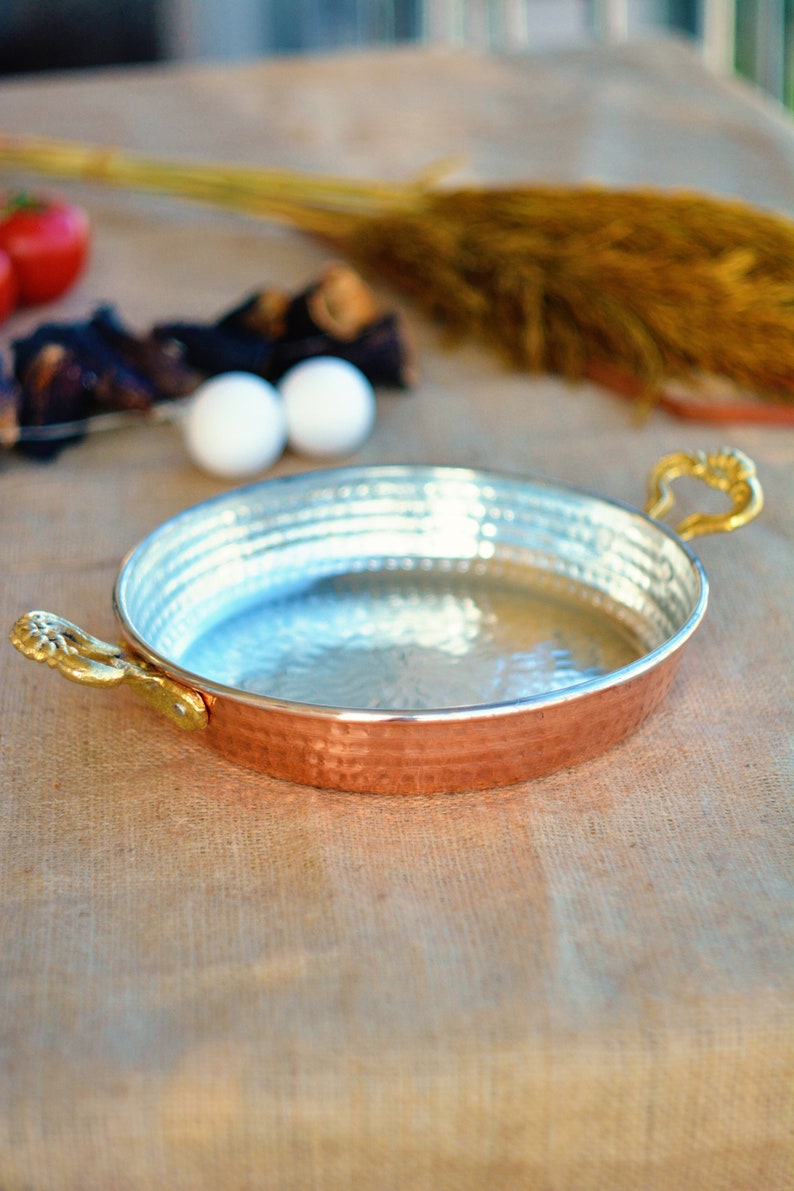 May include: A copper serving dish with two gold handles. The dish has a hammered finish and is sitting on a burlap surface.