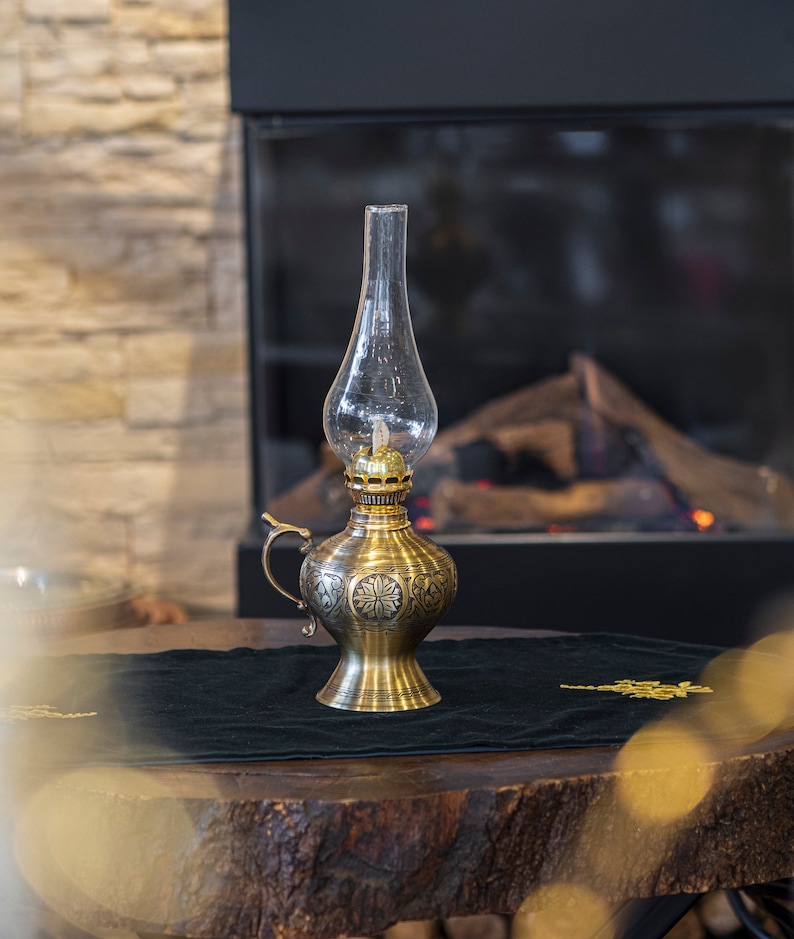 May include: A gold oil lamp with intricate floral designs sits on a wooden table with a black velvet runner. The lamp has a clear glass chimney and a round, bulbous base.
