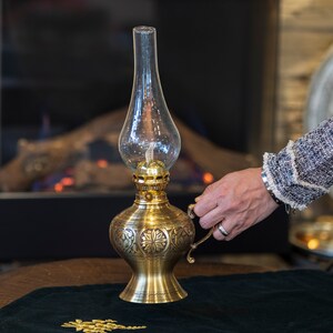 May include: A gold oil lamp with intricate floral designs sits on a dark green velvet surface. The lamp has a clear glass chimney and a round, ornate base.
