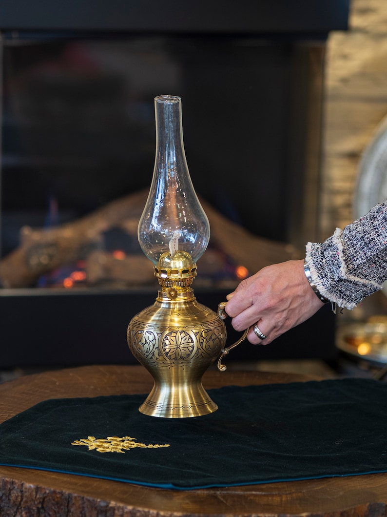May include: A gold oil lamp with intricate floral designs sits on a dark green velvet surface. The lamp has a clear glass chimney and a round, ornate base.