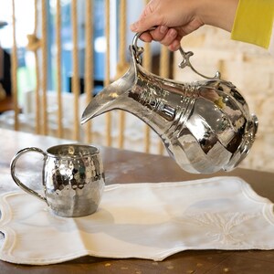 Puede incluir: Una jarra de plata con un diseño ornamentado se está vertiendo en una taza de plata martillada. La jarra y la taza están sobre un mantel individual de lino blanco con un diseño bordado.