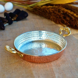 May include: A hammered copper pan with two brass handles. The pan has a shiny silver interior and is sitting on a burlap surface.