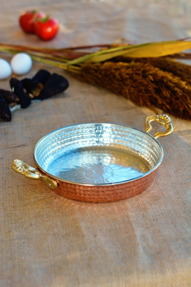 May include: A hammered copper pan with two brass handles. The pan has a shiny silver interior and is sitting on a burlap surface.