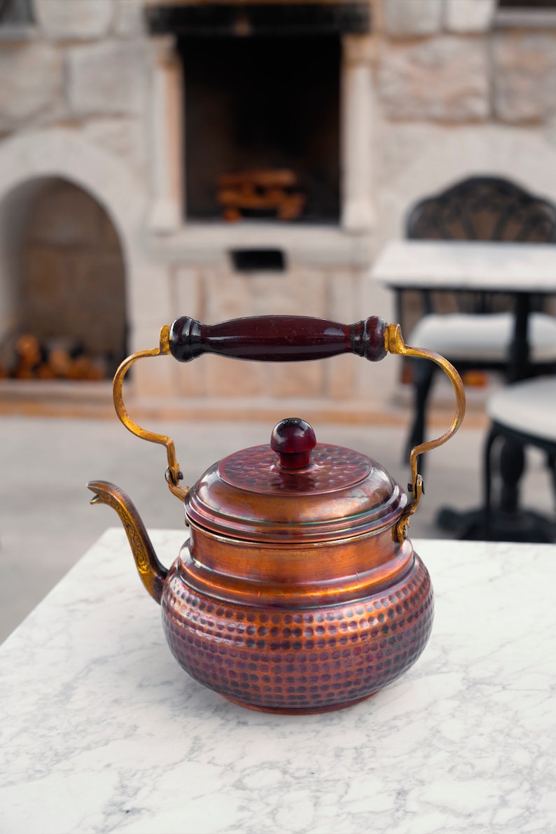 May include: A copper teapot with a hammered finish and a wooden handle. The teapot has a gold-colored spout and lid.