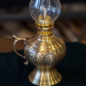 May include: A gold oil lamp with an ornate design and a clear glass chimney. The lamp has a handle and a round base.