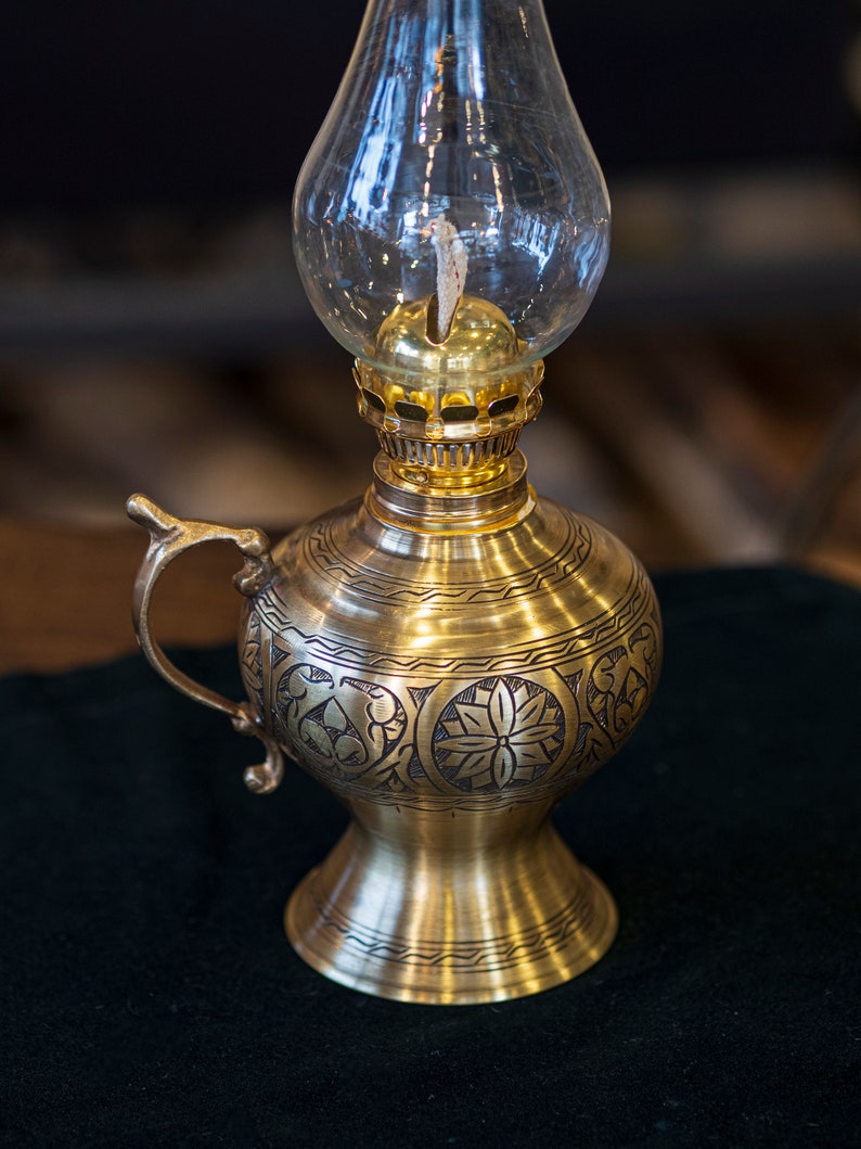 May include: A gold oil lamp with an ornate design and a clear glass chimney. The lamp has a handle and a round base.