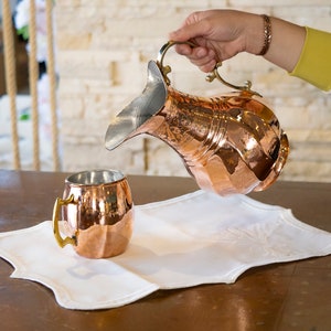 May include: A copper pitcher with a hammered finish and ornate design is being poured into a copper mug with a brass handle. The pitcher and mug are sitting on a white linen placemat.