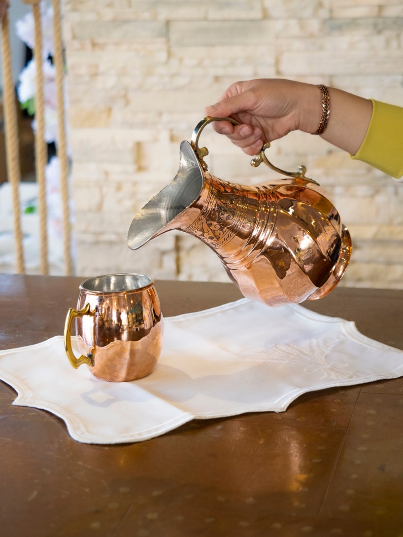 May include: A copper pitcher with a hammered finish and ornate design is being poured into a copper mug with a brass handle. The pitcher and mug are sitting on a white linen placemat.