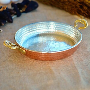 May include: A round copper pan with hammered texture and a shiny silver interior. The pan has two gold handles.