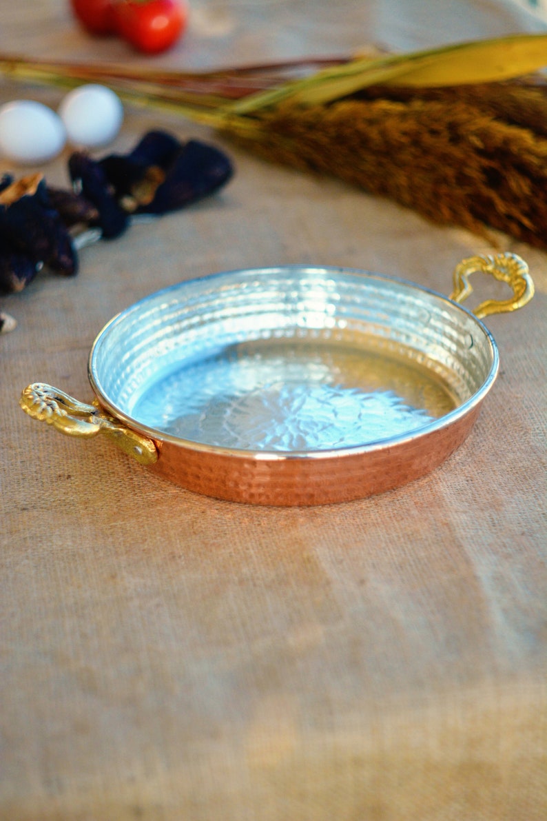 May include: A round copper pan with hammered texture and a shiny silver interior. The pan has two gold handles.