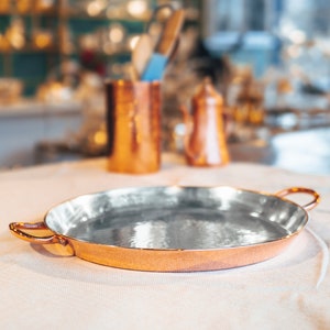 May include: A round copper serving tray with two handles. The tray has a shiny, reflective surface.