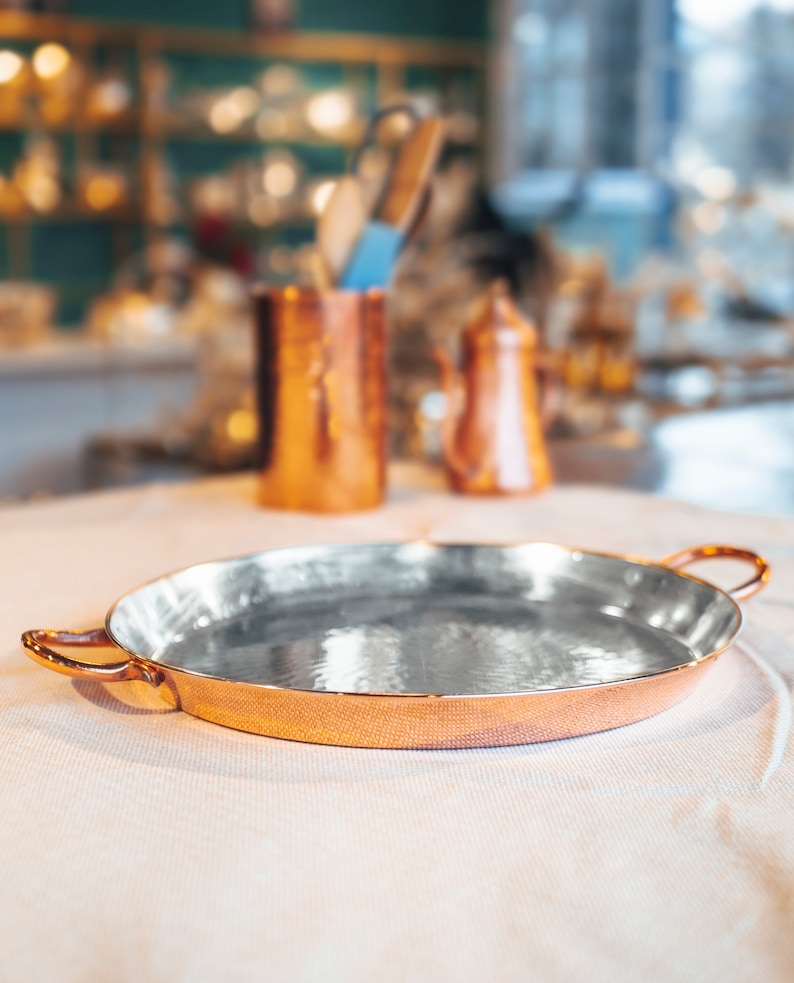 May include: A round copper serving tray with two handles. The tray has a shiny, reflective surface.