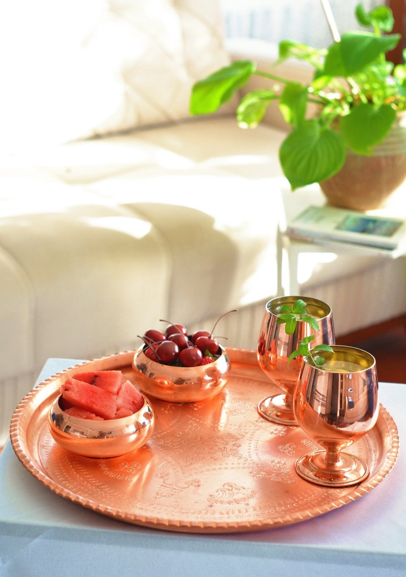 May include: A copper serving tray with a decorative embossed design. The tray holds two copper glasses, a bowl of cherries, and a bowl of watermelon.