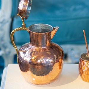 Pu&ograve; includere: Una caraffa in rame martellato con manico e coperchio in ottone. La caraffa ha un corpo rotondo e un'ampia apertura. Il coperchio &egrave; aperto e la caraffa &egrave; appoggiata su una superficie bianca.
