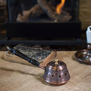 May include: A copper incense burner with a black wooden handle. The burner has an intricate design and is sitting on a hessian surface.
