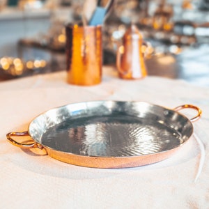 May include: A round copper serving tray with two handles. The tray has a hammered finish and is empty.