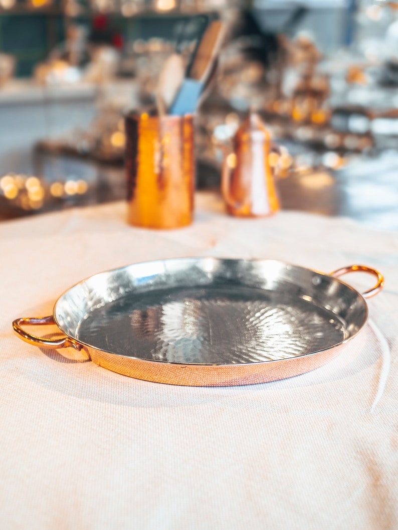 May include: A round copper serving tray with two handles. The tray has a hammered finish and is empty.