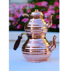May include: A traditional Turkish copper teapot with two tiers and wooden handles. The teapot is polished to a shiny finish and has a decorative design.