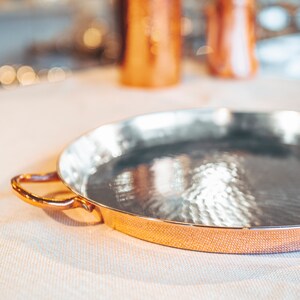 May include: A copper serving tray with a hammered finish and a rounded rim. The tray has a single handle on the left side.