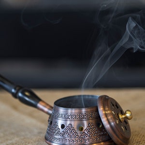 May include: A copper incense burner with a decorative design and a lid. Smoke is rising from the burner, which is sitting on a hessian surface.