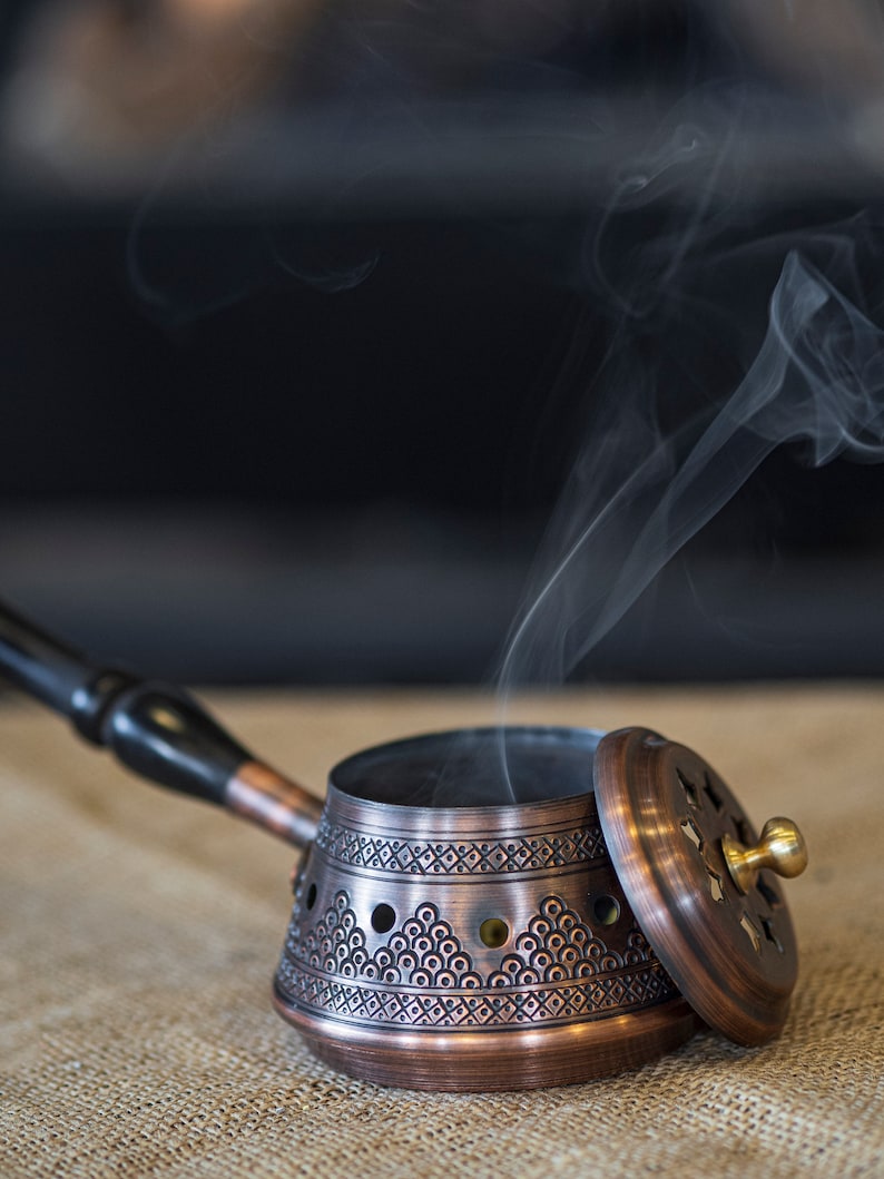 May include: A copper incense burner with a decorative design and a lid. Smoke is rising from the burner, which is sitting on a hessian surface.