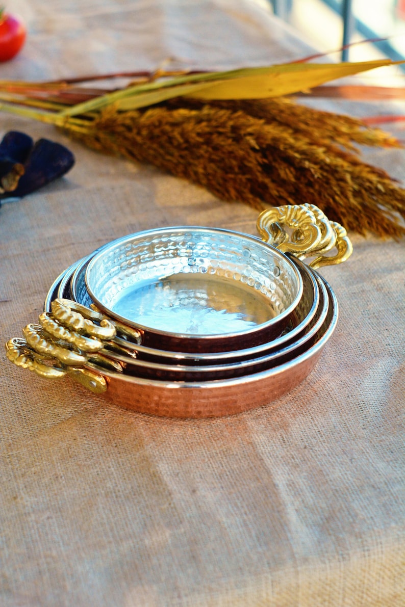 May include: A set of three copper cookware bowls with hammered surfaces and gold-toned handles. The bowls are nested together and have a shiny finish.