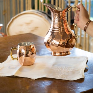 May include: A copper pitcher with an ornate design and a copper mug on a white linen placemat. The pitcher has a handle and a spout. The mug has a handle. The pitcher and mug are on a wooden table.