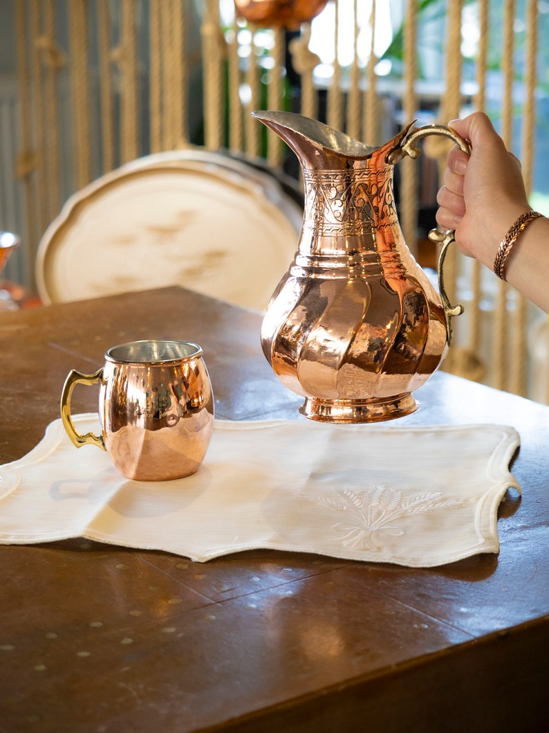 May include: A copper pitcher with an ornate design and a copper mug on a white linen placemat. The pitcher has a handle and a spout. The mug has a handle. The pitcher and mug are on a wooden table.