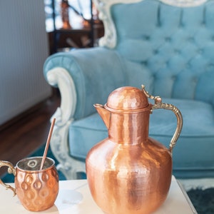 May include: A hammered copper pitcher and mug with a brass handle. The pitcher has a rounded body and a lid. The mug has a rounded body and a handle. The pitcher and mug are sitting on a white table.
