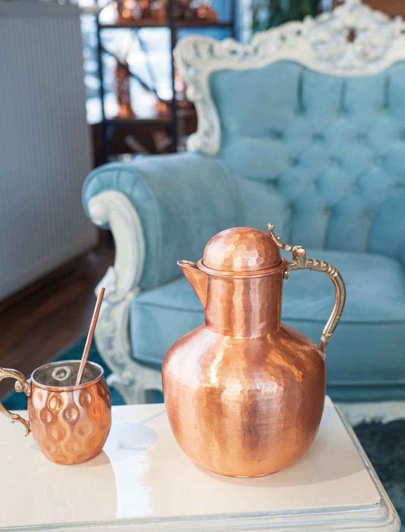 May include: A hammered copper pitcher and mug with a brass handle. The pitcher has a rounded body and a lid. The mug has a rounded body and a handle. The pitcher and mug are sitting on a white table.