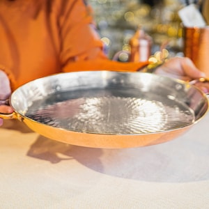 May include: A copper serving dish with a hammered finish and gold-coloured handles. The dish is empty and is being held by a person's hands.