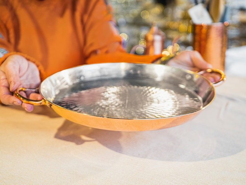 May include: A copper serving dish with a hammered finish and gold-coloured handles. The dish is empty and is being held by a person's hands.