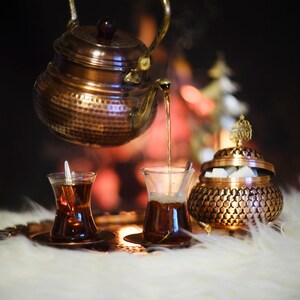 May include: A copper teapot pours tea into two traditional Turkish tea glasses on a tray with a copper sugar bowl. The glasses are filled with dark tea and the sugar bowl is filled with white sugar cubes.