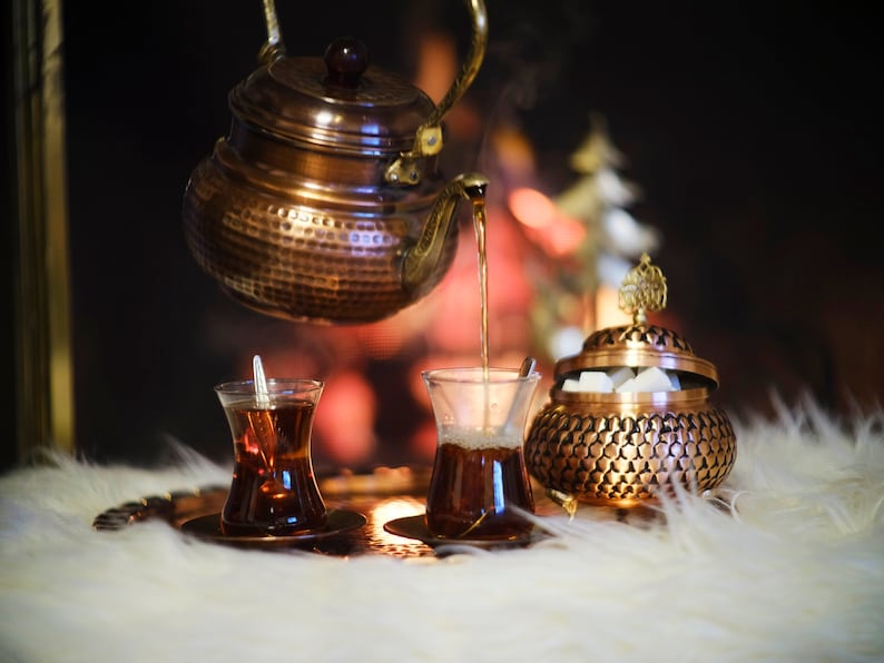 May include: A copper teapot pours tea into two traditional Turkish tea glasses on a tray with a copper sugar bowl. The glasses are filled with dark tea and the sugar bowl is filled with white sugar cubes.