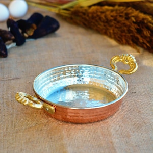 May include: A small, round copper pan with a hammered finish and two gold handles. The pan is empty and sitting on a burlap surface.