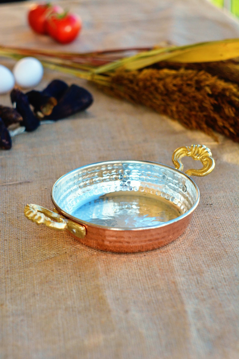May include: A small, round copper pan with a hammered finish and two gold handles. The pan is empty and sitting on a burlap surface.