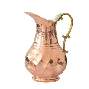 May include: A copper pitcher with a hammered finish and an ornate design around the neck. The pitcher has a brass handle and a rounded base.