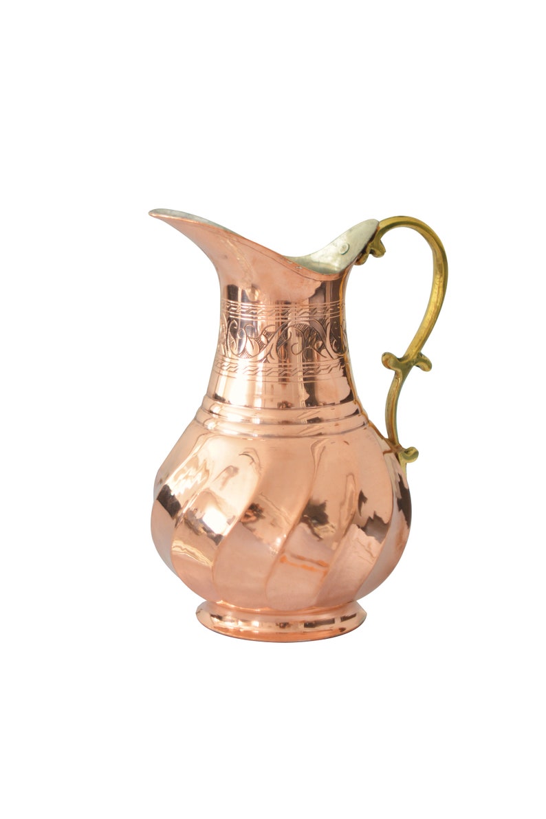 May include: A copper pitcher with a hammered finish and an ornate design around the neck. The pitcher has a brass handle and a rounded base.