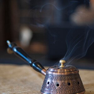 May include: A copper incense burner with a black wooden handle and intricate designs. Smoke rises from the burner, which is sitting on a hessian surface.