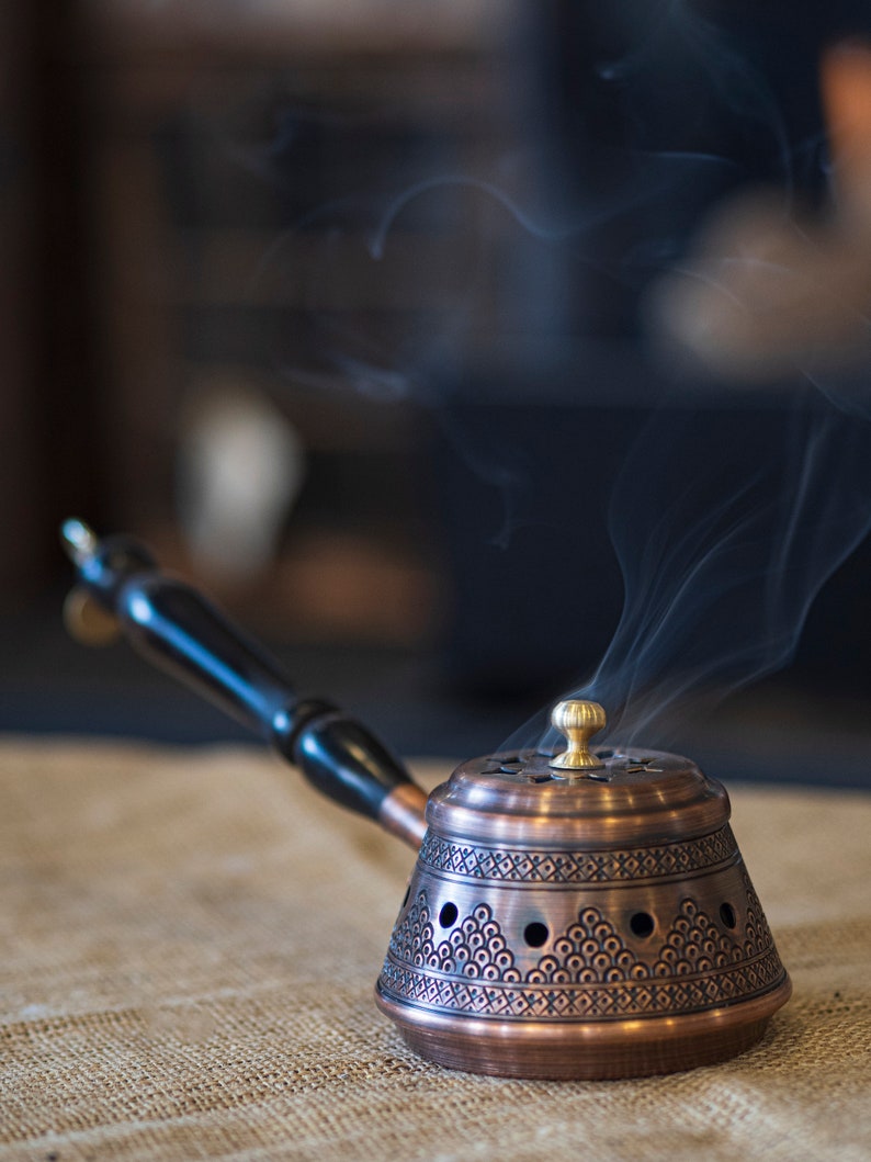 May include: A copper incense burner with a black wooden handle and intricate designs. Smoke rises from the burner, which is sitting on a hessian surface.